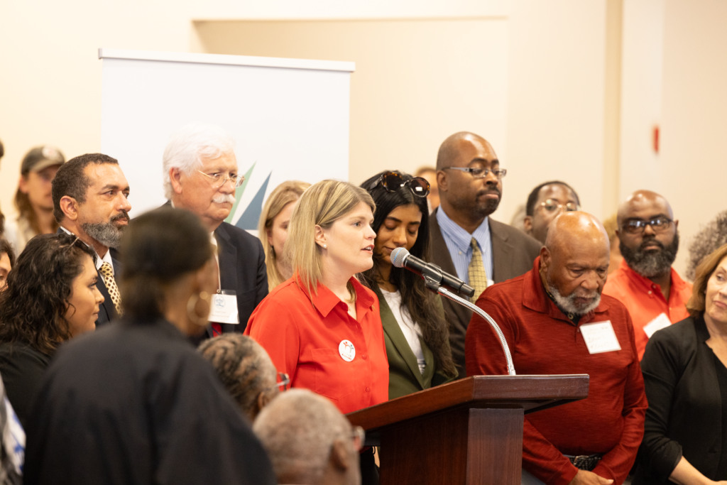 A white woman with blonde hair speaks while standing at a lectern with several people behind her.