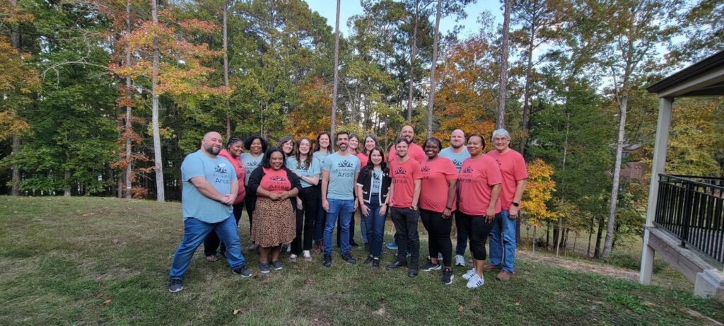 A group of 17 people smile for a photo while standing on a green hilltop. All are wearing red or green T-shirts with the Alabama Arise logo. Behind them is a line of evergreen trees and trees with leaves turning yellow or orange for fall.