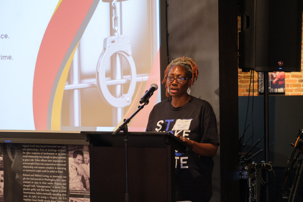 A Black woman wearing glasses and a black T-shirt speaks behind a black lectern. Behind her is a screen with an image of handcuffs in front of jail bars.